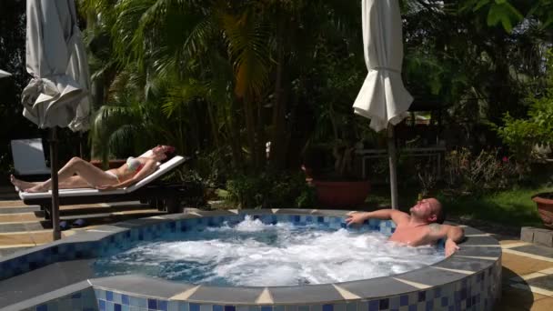 Uma mulher está deitada numa espreguiçadeira e bronzeada. Um homem descansa no jacuzzi — Vídeo de Stock