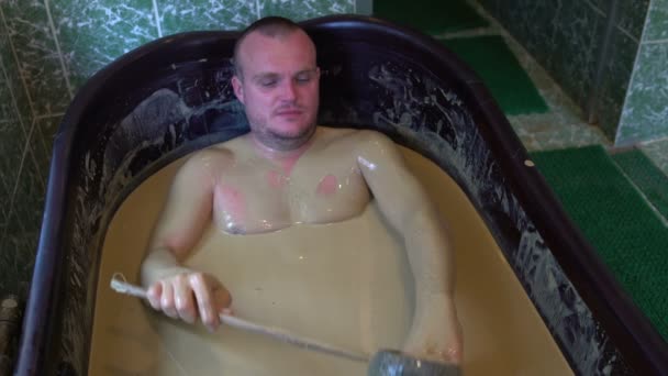 El hombre yace en un baño con barro curativo y se derrama fuera del cubo — Vídeos de Stock