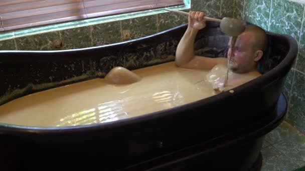 El hombre yace en el baño de barro y derrama suciedad sobre su cabeza fuera del cubo — Vídeo de stock