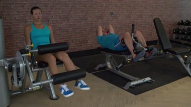 Kadın bir simülatör üzerinde bacaklarını sallıyor. Adam basın eğimli bir bankta sallıyor.