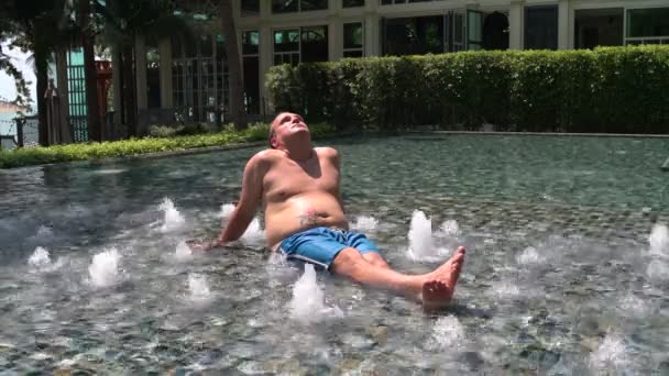 Человек сидит в неглубоком бассейне и загорает — стоковое видео