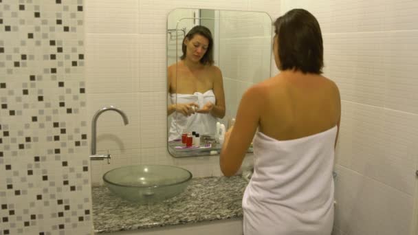 Een vrouw in een handdoek zet een vochtinbrengende crème op haar schouders voor een badkamerspiegel — Stockvideo