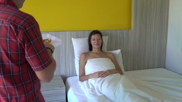 Una mujer está acostada en la cama. Un hombre trae a una mujer un waffle y café en una bandeja y la besa — Vídeo de stock