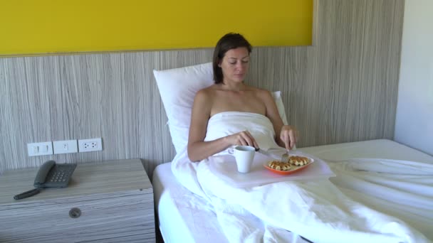 Жінка їсть вафлі з ножем і виделкою, лежачи в ліжку в готельному номері — стокове відео