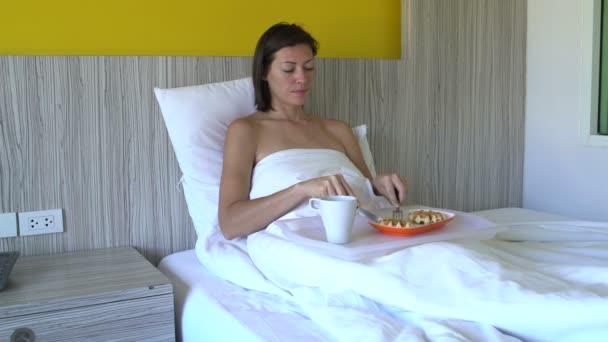 Женщина завтракает с вафлями и пьет кофе, лежащий в постели в номере отеля — стоковое видео