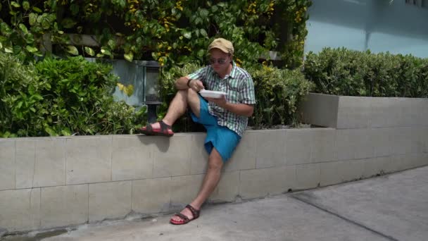 Уличная еда. Человек, сидящий на улице на парапете и поедающий тушеные овощи из контейнера — стоковое видео