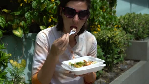 Streetfood. eine Frau, die auf der Straße auf einer Brüstung sitzt und Gemüse aus einem Container isst — Stockvideo