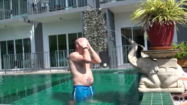 En man står i poolen under strålen av vatten som rinner ur stammen av en sten elefant — Stockvideo