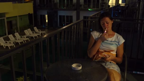 Une femme s'assoit avec un smartphone à une table sur le balcon dans une chambre d'hôtel et fume — Video