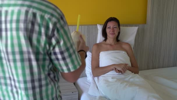Un hombre trae un coco joven con una paja a una mujer en la cama. Mujer bebiendo leche de coco — Vídeo de stock