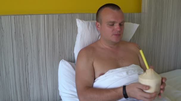 Homem na cama bebendo através de um tubo de leite de coco de um jovem coco — Vídeo de Stock