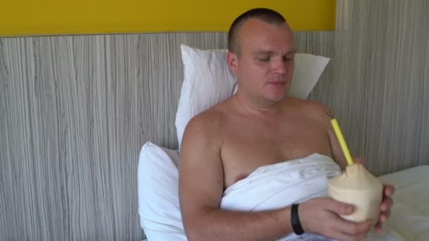 Mann im Bett trinkt Kokosmilch aus einer jungen Kokosnuss — Stockvideo