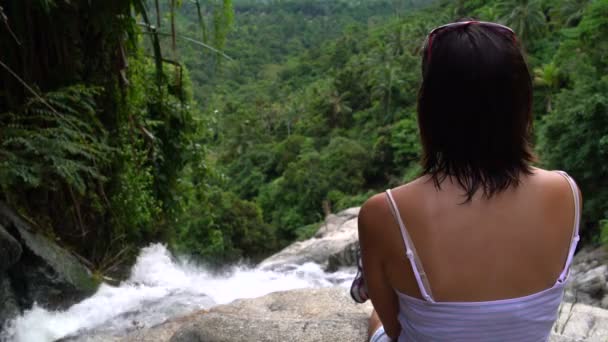 La mujer se sienta en la parte superior de la cascada y mira hacia abajo — Vídeo de stock