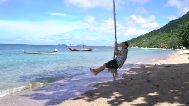 Чоловік в сонцезахисних окулярах гойдається на мотузці на пляжі на узбережжі — стокове відео