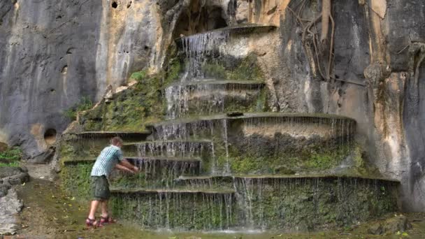 Человек приближается к искусственному каскадному водопаду в скале и умывается — стоковое видео