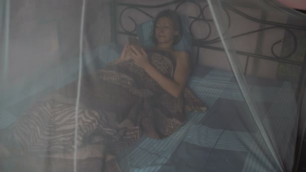 Μια γυναίκα βρίσκεται στο κρεβάτι με έναν θόλο και χρησιμοποιεί ένα smartphone — Αρχείο Βίντεο