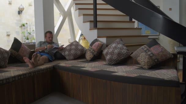En man läser en tidskrift som liggande på soffan hemma — Stockvideo