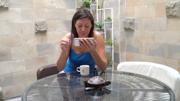 Vrouw drinken thee en kijken naar grappige video's op de telefoon zittend aan een tafel — Stockvideo