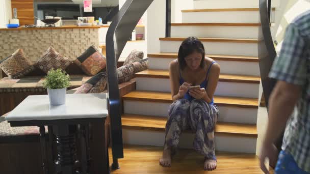 Женщина сидит на лестнице в доме и пользуется смартфоном. Мужчина поднимается по лестнице — стоковое видео
