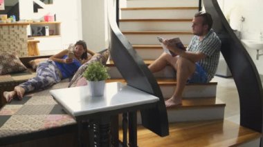 Bir adam evde merdivenlerde oturan bir kitap okuyor. Bir kadın kanepede yatıyor ve bir akıllı telefon kullanıyor