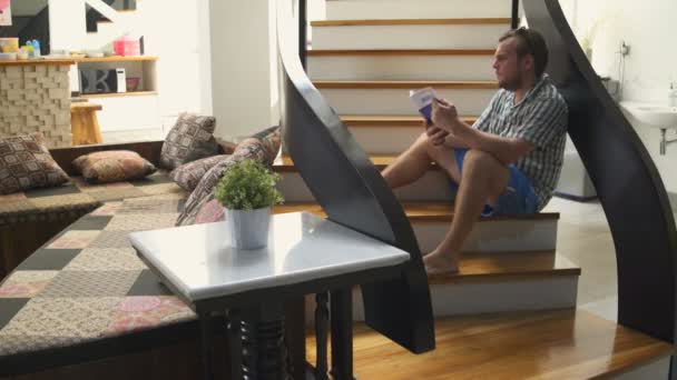 Ein Mann sitzt auf der Treppe im Haus und liest ein Buch — Stockvideo