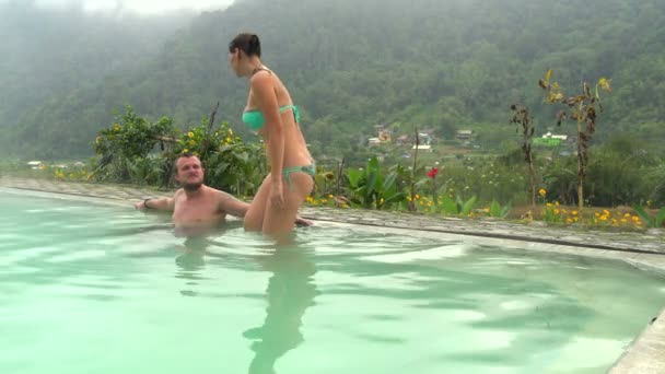 Человек сидит в бассейне с термальной водой. Женщина входит в бассейн и целует мужчину — стоковое видео