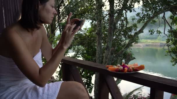 Uma mulher em uma toalha tira fotos de um prato de frutas contra o lago — Vídeo de Stock