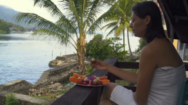Mujer comiendo una fruta larga sentada en una toalla en el fondo de un lago y montañas — Vídeos de Stock