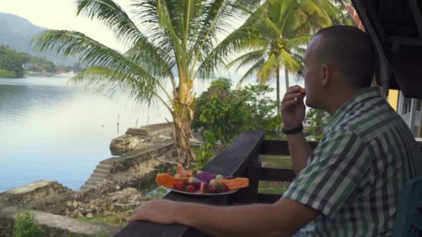 Een man eet papaya en kijkt naar het meer en de bergen — Stockvideo