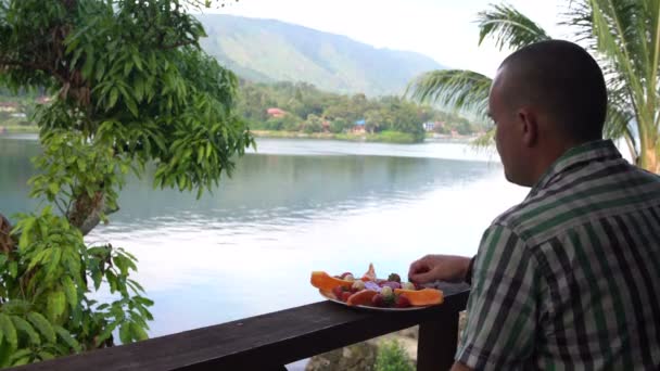 Un hombre come fresas y mira el lago y las montañas — Vídeos de Stock