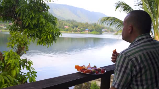 Человек ест фрукты и смотрит на озеро и горы — стоковое видео