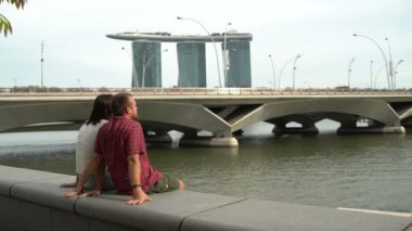 Adam ve kadın bir parapetin üzerinde Marina Bay Sands Hotel bakan nehir dolgu üzerinde oturuyor