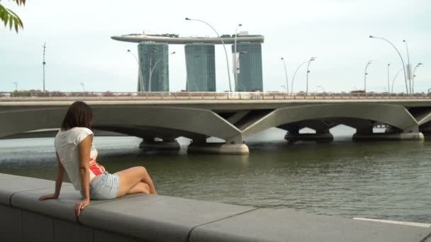 Mujer se sienta en un parapeto en el terraplén del río con vistas al Marina Bay Sands Hotel en Singapur — Vídeos de Stock