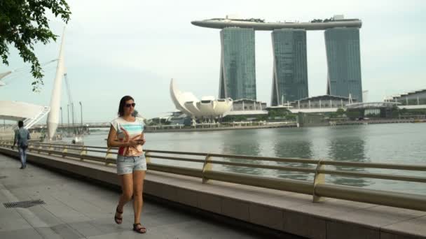 Жінка прогулянках уздовж набережної річки і робить знімки Marina Bay Sands Hotel в Сінгапурі — стокове відео