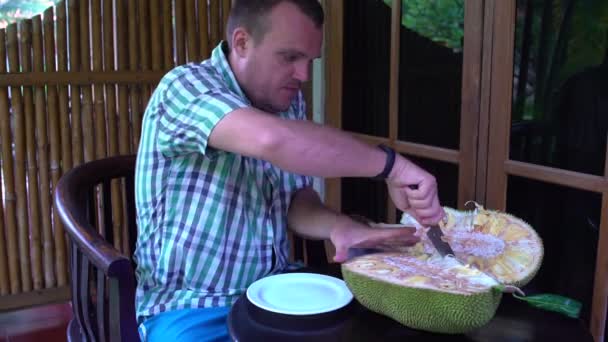 Egy férfi csökkenti a kenyérfa gyümölcse egy késsel a teraszon — Stock videók