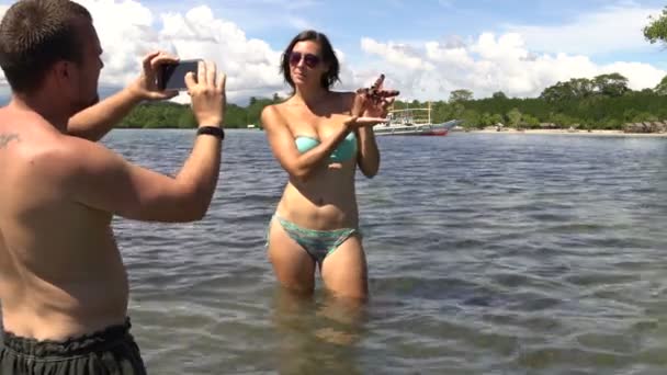 En man tar bilder en kvinna i en baddräkt står i havet med en stjärna i händerna — Stockvideo
