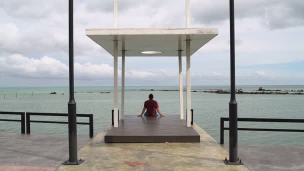 Ein Mann sitzt auf einem Steg und blickt auf das Meer — Stockvideo