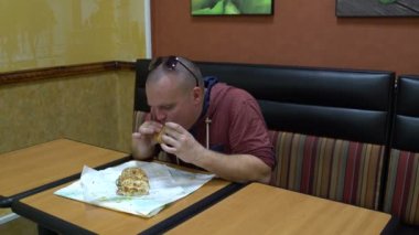 Adam bir kafede bir masada oturur ve bir sandviç yiyor.