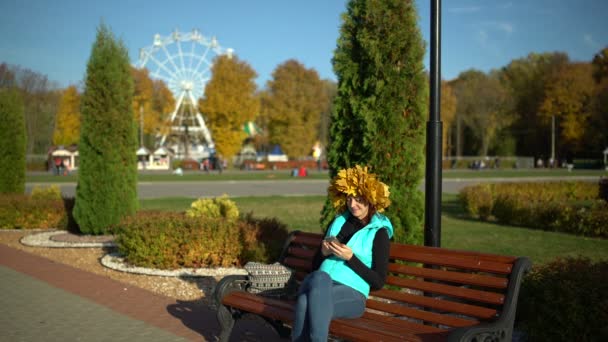 Una mujer se sienta en un banco en el parque y usa un teléfono inteligente — Vídeo de stock