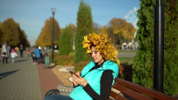 Vrouw met een kroon van de esdoorn op haar hoofd zit op een bankje — Stockvideo