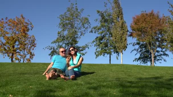 Donna e uomo sedersi nel parco sul prato a piedi nudi e utilizzare uno smartphone — Video Stock