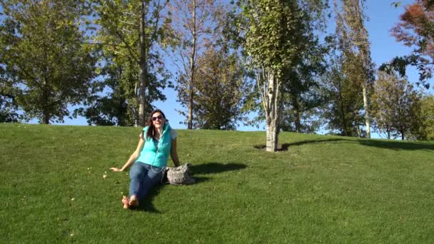 Женщина сидит в парке на лужайке босиком и разговаривает на гарнитуре — стоковое видео