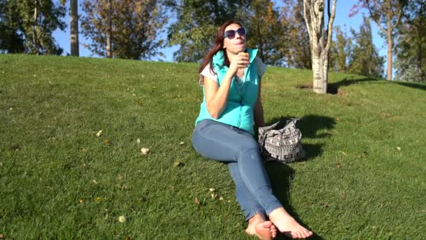 Woman sits in the park on the lawn barefoot and eats patty — Stock Video