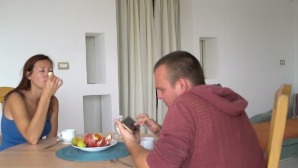 Pareja sentada en la mesa y hablando. Un hombre usa un teléfono inteligente. Mujer comiendo plátano — Vídeo de stock