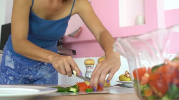 Een vrouw met een mes snijdt een komkommer en een tomaat op een snijplank op een tafel in de keuken — Stockvideo