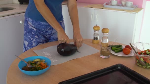 Vrouw snijdt aubergine met een mes op een snijplank op een tafel in de keuken — Stockvideo