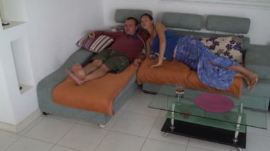 Bir adam kanepede yatıyor ve Tv izliyor. Bir kadın gelir ve ileri yatıyor