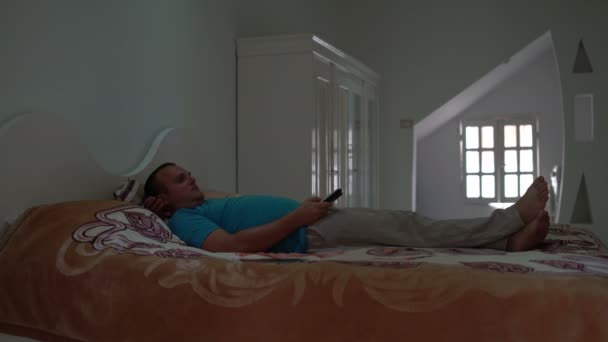 Чоловік лежить у спальні на ліжку з пультом і дивиться телевізор — стокове відео