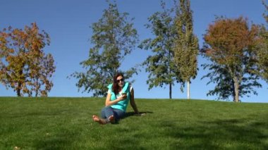 Kadın parkta yeşil çimenlerin üzerinde yalınayak oturur ve smartphone kullanır