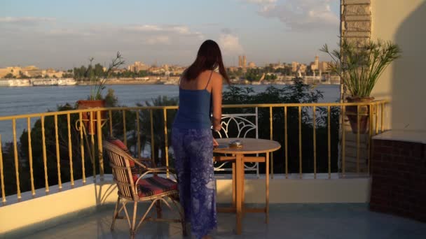 Uma mulher chega ao terraço com vista para o rio e senta-se à mesa . — Vídeo de Stock